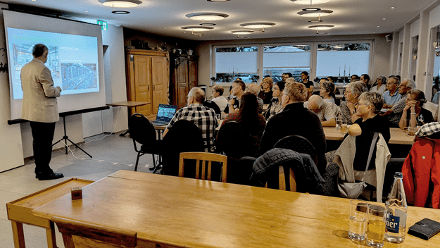 Informationsabend zum Thema künstliche Intelligenz im Restaurant Nollen. Referent Christian Blattner präsentiert vor über 30 Teilnehmenden, die an mehreren Tischen aufmerksam zuhören. Auf der Leinwand ist eine Präsentation zum Thema künstliche Intelligenz zu sehen.