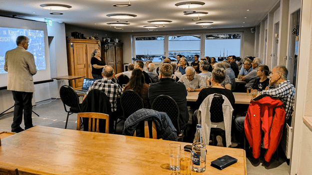 Informationsabend zum Thema künstliche Intelligenz im Restaurant Nollen. Referent Christian Blattner präsentiert vor über 30 Teilnehmenden, die an mehreren Tischen aufmerksam zuhören. Auf der Leinwand ist eine Präsentation zum Thema künstliche Intelligenz zu sehen.
